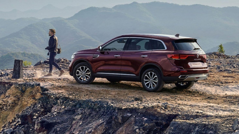 SUV Renault Koleos color vino tinto estacionada en terreno rocoso con montañas de fondo.